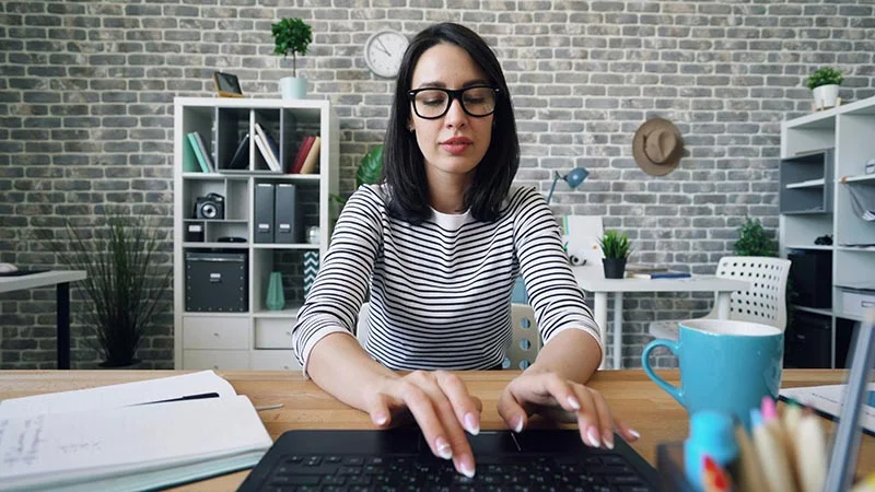 Junge Frau mit Brille sitzt in einem modernen Büro am Schreibtisch und tippt konzentriert auf ihrem Laptop.