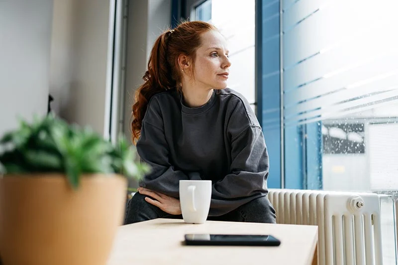Junge Frau sitzt neben dem Fenster an einem kleinen Tisch und schaut melancholisch nach draußen in den Regen.